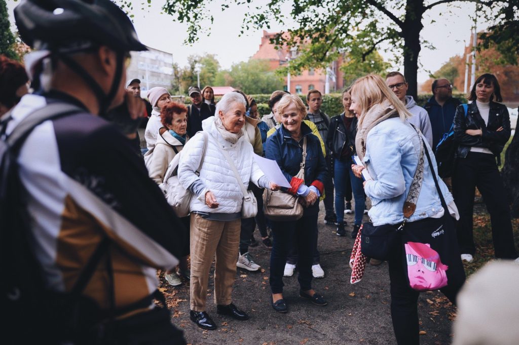 Kolorowa, pozioma fotografia: grupa osób w różnym wieku zebrała się na skwerze. Trwa spacer po osiedlu. Na środku zdjęcia starsza kobieta opowiada historię, pokazując coś na wydrukowanych zdjęciach.
