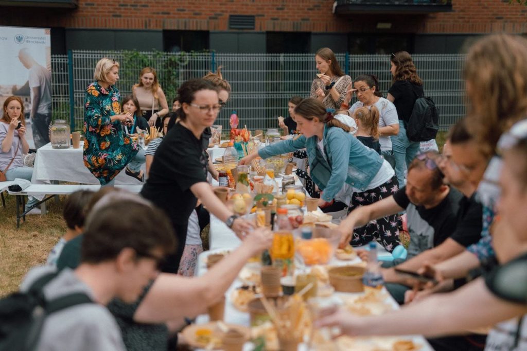 Kolorowa, pozioma fotografia: grupa osób w różnym wieku zebrała się wokół długiego stołu pełnego owoców, ciast i innych przekąsek. Trwa sąsiedzki piknik.