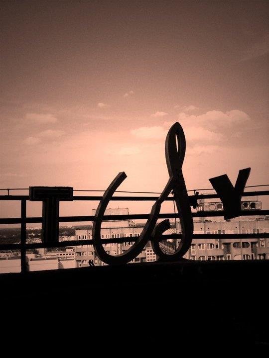 Zdjęcie: duże litery T, S i Y przyczepione do barierek. W tle pomarańczowe niebo i szczyty wieżowców.