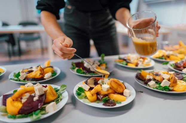 Zbliżenie na rozłożone w dwóch rzędach talerze z nałożonymi porcjami pięknie wyglądającej kolorowej sałatki. Nad talerzami pochyla się młody człowiek i polewa je delikatnie sosem.
