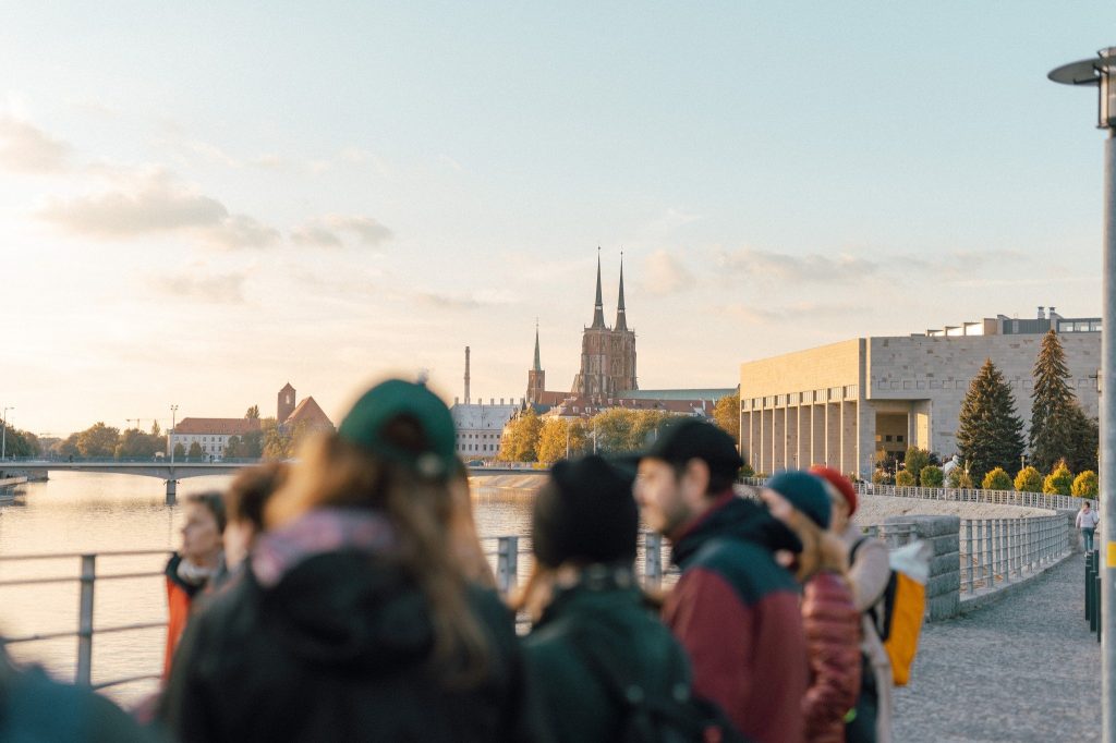 Kolorowa, pozioma fotografia: na pierwszym planie niewyraźna grupa osób w różnym wieku słuchająca opowieści przewodnika spaceru. W tle widoczna Odra oraz fragment nabrzeża z budynkiem biblioteki uniwersyteckiej. Dalej widoczne wieże Katedry.