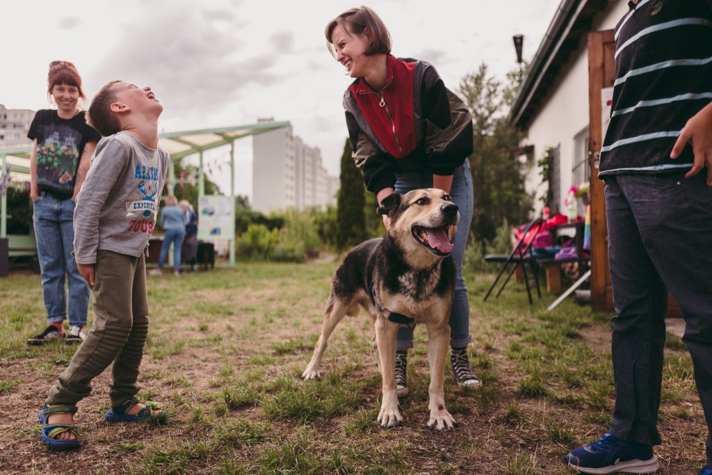 Kolorowa, pozioma fotografia przedstawiająca kobietę, głaszczącą psa oraz dziecko na terenie ogródka działkowego. Oboje śmieją się.