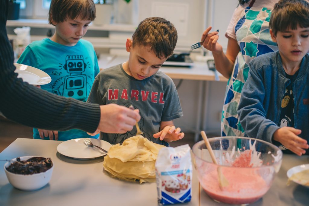 Kolorowa, pozioma fotografia. Trójka kilkuletnich chłopców stoi przy stole i dekorują naleśniki. Na stole stoi talerz z naleśnikami, miska z kremem czekoladowym oraz duża miska z różową polewą.