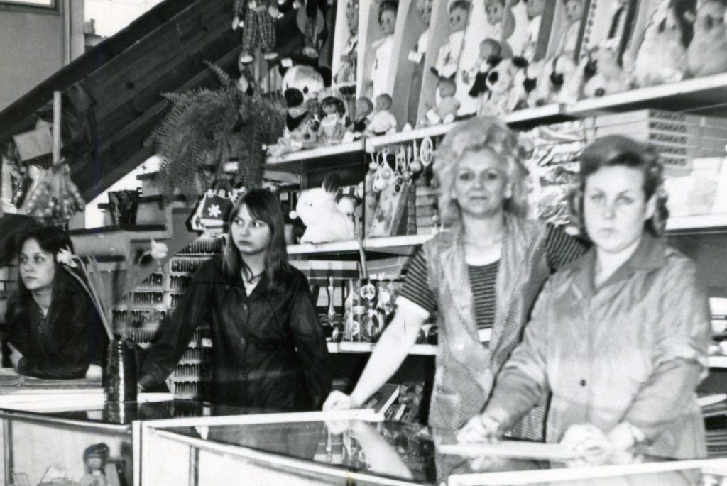 Archiwalne, czarno-białe zdjęcie. Ekspedientki na tle półek sklepowych, na których stoją zabawki.