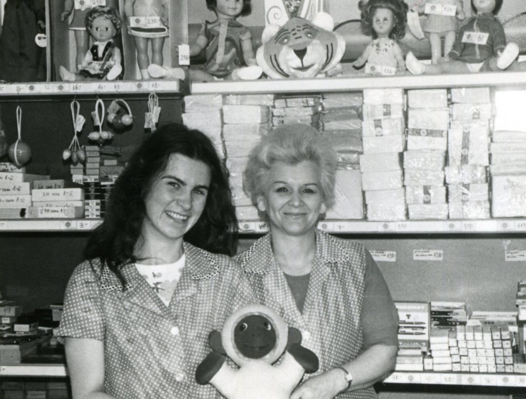 Archiwalne, czarno-białe zdjęcie. Dwie ekspedientki w kraciastych fartuszkach pozują na tle półek sklepowych, na których stoją zabawki.