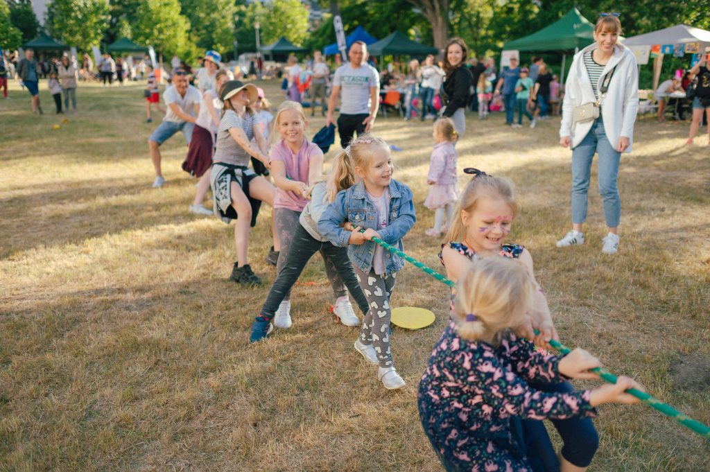 Kolorowa, pozioma fotografia przedstawiająca grupę dzieci i dorosłych bawiących się w przeciąganie liny. Trwa sąsiedzki piknik na łące.