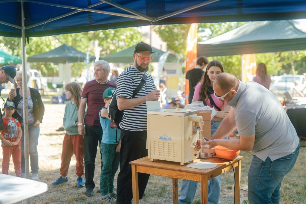 zdjęcie: Marcin Szczygieł Kolorowa, pozioma fotografia przedstawia długą kolejkę osób w różnym wieku czekających przed stanowiskiem z tradycyjnymi rurkami z kremem.