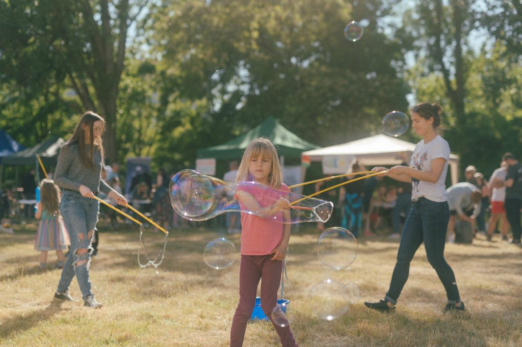 zdjęcie: Marcin Szczygieł Kolorowa, pozioma fotografia przedstawia kilkuletnią dziewczynkę puszczającą duże bańki mydlane podczas pikniku na łące.