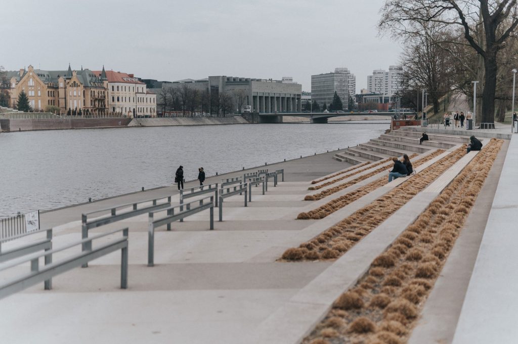 Kolorowa, pozioma fotografia. Widok na nabrzeże, rzekę oraz znajdującą się na drugim brzegu Bibliotekę.