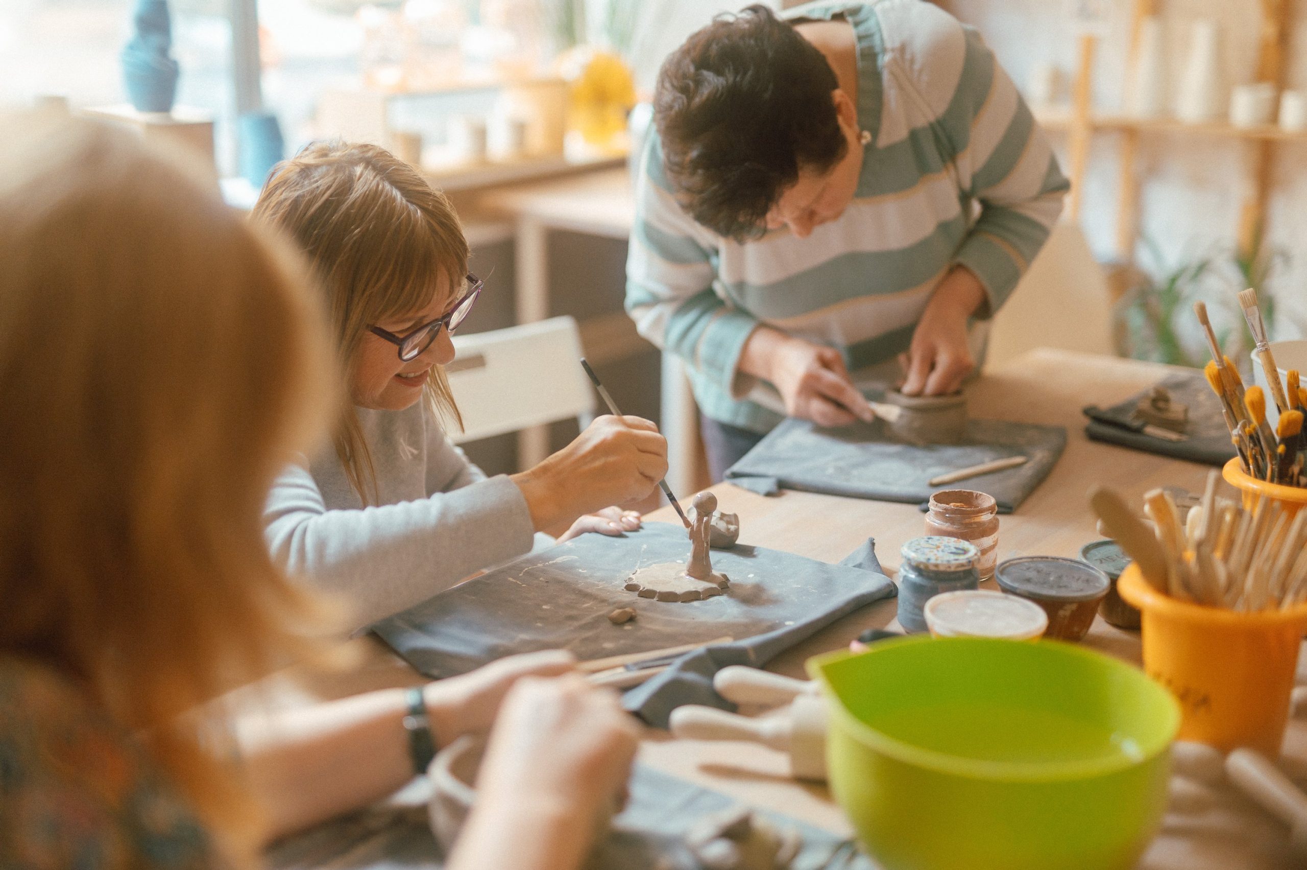 Kolorowa, pozioma fotografia: przy stole warsztatowym w jasnym pomieszczeniu siedzą trzy seniorki, biorą udział w warsztatach ceramicznych. Każda z nich wykonuje z gliny jakiś przedmiot - jedna rzeźbi figurkę, druga tworzy miseczkę, trzecia robi kubek. Na stole leżą też wałki, pędzle, słoiki z farbą i miski z wodą.
