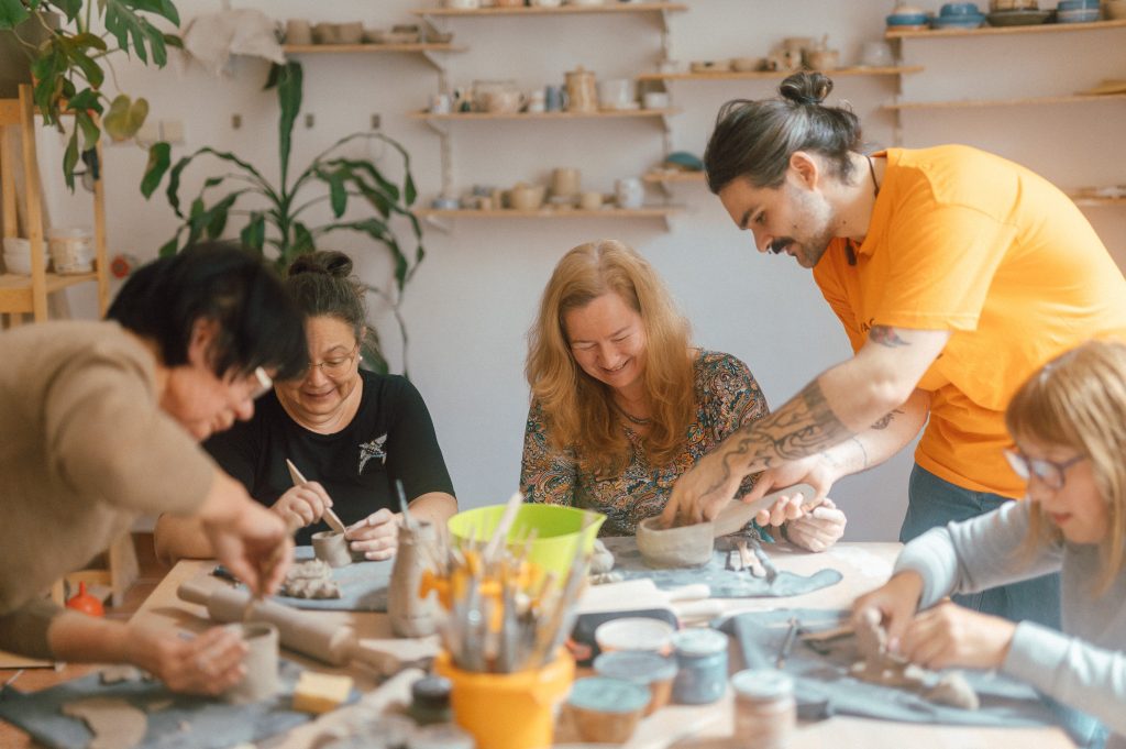 Kolorowa, pozioma fotografia: pięć seniorek siedzi przy dużym stole i rzeźbi w glinie. Młody mężczyzna pomaga jedne z nich przykleić ucho do kubka.