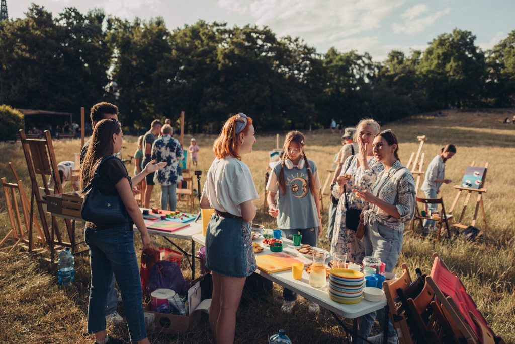 Kolorowa, pozioma fotografia przedstawiająca grupę osób w różnym wieku zebraną na łące w parku. Wszędzie rozstawione są sztalugi i obrazy. W centrum zdjęcia stół z przekąskami/