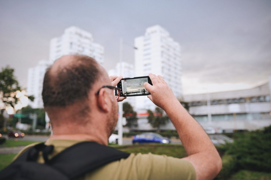 Kolorowa, pozioma fotografia. Na pierwszym planie mężczyzna robiący zdjęcie charakterystycznym wieżowcom na Placu Grunwaldzkim.