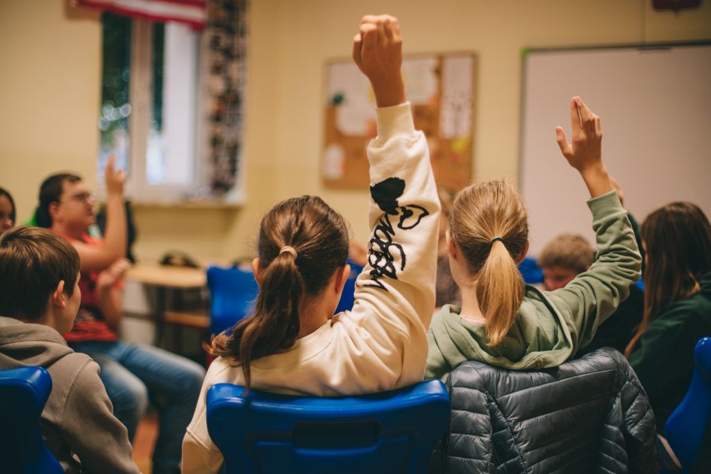 Kolorowa, pozioma fotografia. Zdjęcie wykonane w niedużej klasie. Grupa uczniów i uczennic siedzi na krzesłach ustawionych w okrąg. Część z nich zgłasza się poprzez podniesienie ręki.