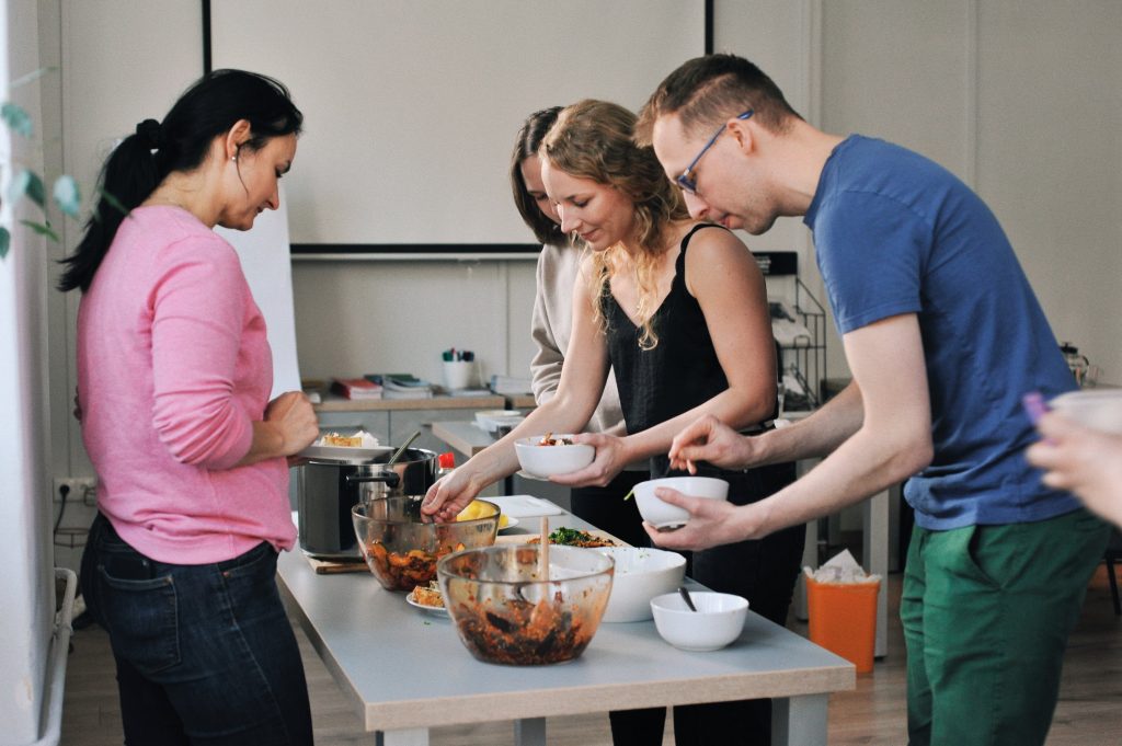 zdjęcie: Anna Pazdej Kolorowa, pozioma fotografia: przy stole warsztatowym w jasnym pomieszczeniu stoją trzy kobiety i mężczyzna. Wspólnie przygotowują posiłek w ramach warsztatów kulinarnych. Na stole stoją miski z półproduktami, naczynia i garnki.