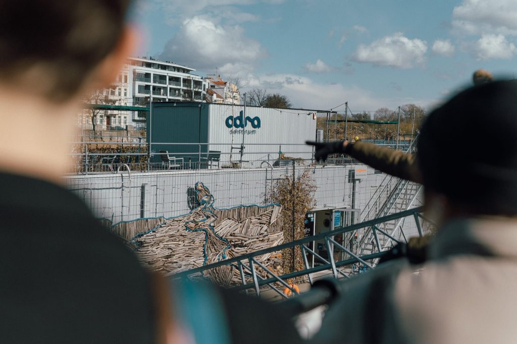 Kolorowa fotografia: osoba poza kadrem, palcem wskazuje budynek na rzece - Odra Centrum.