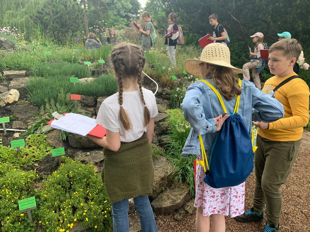 Grupa dzieciaków spaceruje po ogrodzie botanicznym, przy okazji rozwiązując kreatywne zadania.