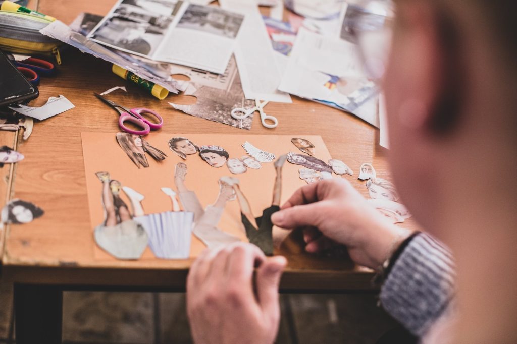 Kolorowa, pozioma fotografia. Zbliżenie na ręce układające kolaż z  fragmentów zdjęć. W tle widoczne porozkładane na stole nożyczki, kleje i gazety.