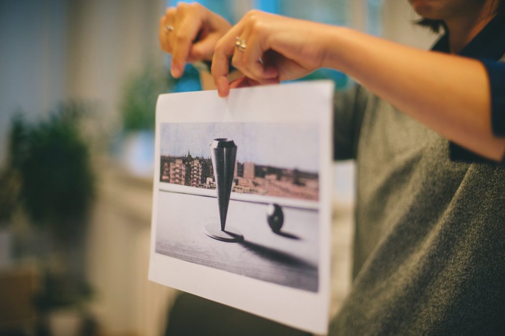 Kolorowa, pozioma fotografia przedstawiająca ręce, które trzymają wydrukowane na kartce zdjęcie rzeźby w kształcie odwróconego stożka.