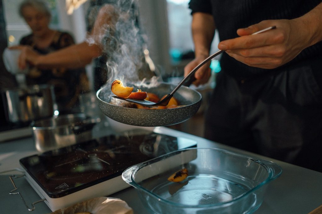 Kolorowa, pozioma fotografia przedstawiająca zbliżenie na dłonie wyjmujące z patelni, jeszcze parujące kawałki brzoskwiń.