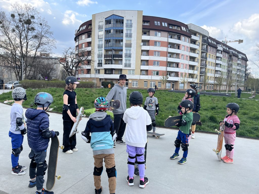 materiały organizatora Kolorowa, pozioma fotografia. Dziesiątka dzieci zebrała się na betonowym chodniku. Każde z nich ma kask i ochraniacze, a w ręce trzyma deskorolkę. Dzieci zebrały się wokół młodego mężczyzny, który poprowadzi warsztaty.
