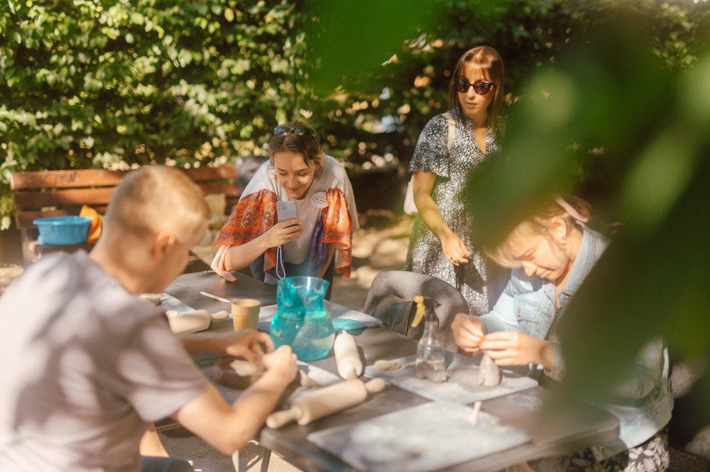 Kolorowa, pozioma fotografia, przedstawiająca warsztaty ceramiczne, odbywające się na świeżym powietrzu. Przy dużym stole Dwie osoby wykonują gliniane rzeźby. Nad nimi pochyla się młoda kobieta i robi im zdjęcie telefonem.