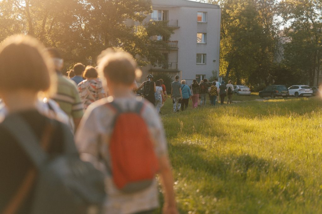 fot. Marcin Szczygieł Kolorowa, pozioma fotografia przedstawiająca grupę osób, spacerujących wąską ścieżką przez łąkę między blokami.