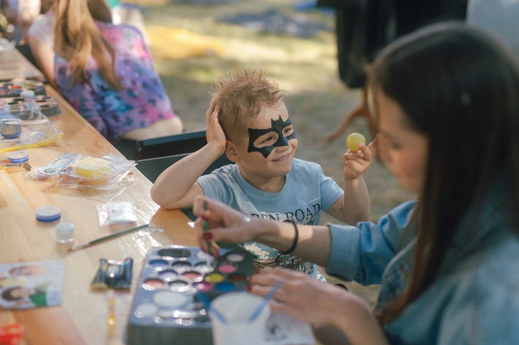 Kolorowa, pozioma fotografia, przedstawiająca kilkuletniego chłopca z namalowaną maską na buzi. Obok niego siedzi animatorka z pędzelkiem w ręku. Przed nimi na długim stole leżą farby w różnych kolorach.