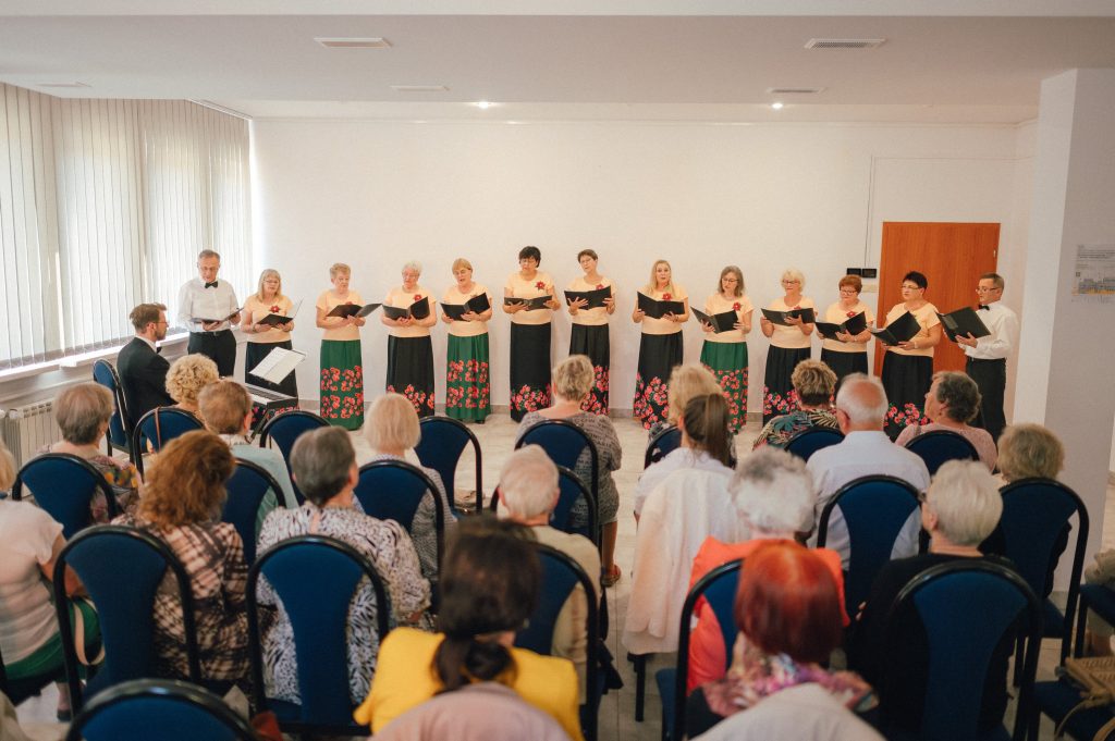 Kolorowa, pozioma fotografia, przedstawiająca występ chóru Grunwald. W niedużym pomieszczeniu występuje 11 seniorek i dwóch seniorów. Po lewej stronie przy keyboardzie siedzi dyrygent chóru. Na pierwszym planie siedząca publiczność.   
