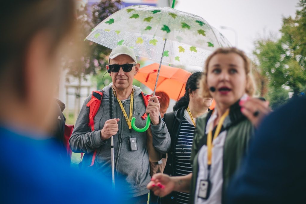 zdjęcie: Marta Sobala Kolorowa, pozioma fotografia przedstawiająca grupę osób na spacerze. Ostrość skupiona jest na starszym mężczyźnie z parasolem. Mężczyzna ma ciemne okulary, w ręce trzyma białą laskę, a w uchu ma słuchawkę. Przed nim widoczna jest przewodniczka, która przez mikrofon opowiada o mijanych miejscach.