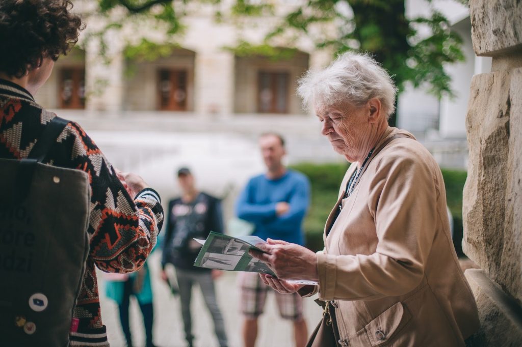 zdjęcie: Marta Sobala Kolorowa, pozioma fotografia przedstawia starszą kobietę czytającą ulotkę. Za kobietą widoczne kolejne osoby. To uczestnicy i uczestniczki spaceru po osiedlu.