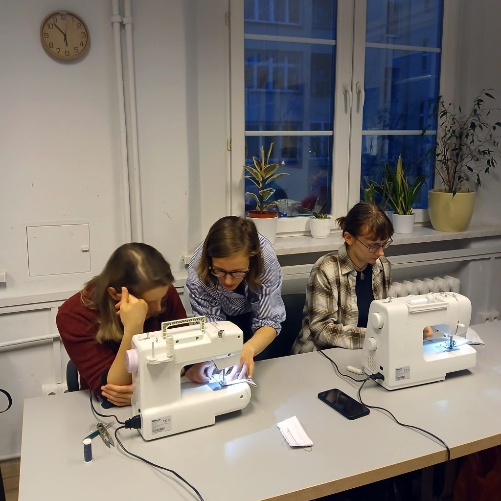 Kolorowa fotografia wykonana podczas warsztatów szycia. Przy długim stole siedzą dwie młode kobiety. Przed nimi stoją dwie maszyny do szycia. Prowadząca zajęcia pochyla się nad jedną z kobiet i pomaga jej z ułożeniem materiału.