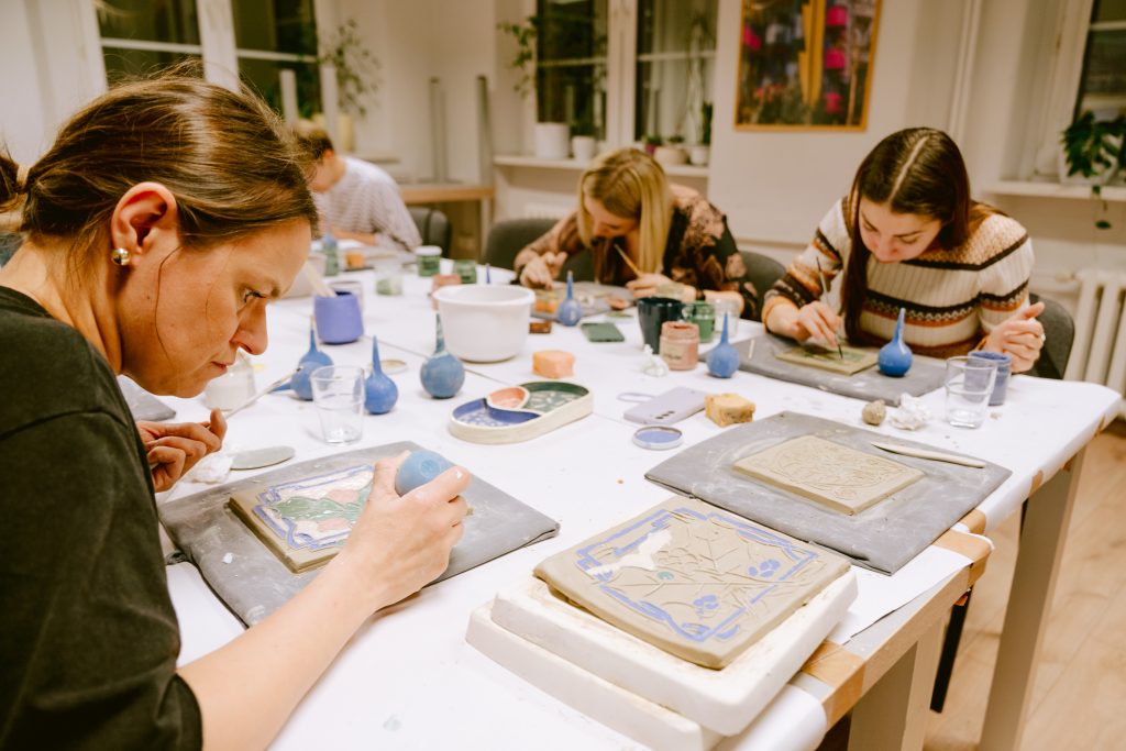 Kolorowa fotografia wykonana podczas warsztatów ceramicznych. W dużym pomieszczeniu przy stole siedzi grupa osób tworzących z gliny kolorowe kafle.