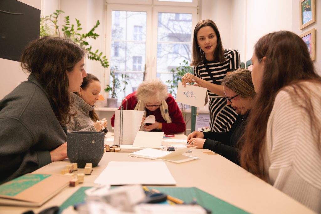 Kolorowa fotografia wykonana podczas warsztatów. Przy stole siedzi grupa osób tworzących grafiki przy pomocy pieczątek i kolorowych tuszy.