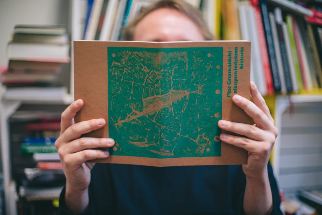 Kolorowa fotografia przedstawia zbliżenie na mężczyznę trzymającego zin na wysokości twarzy.