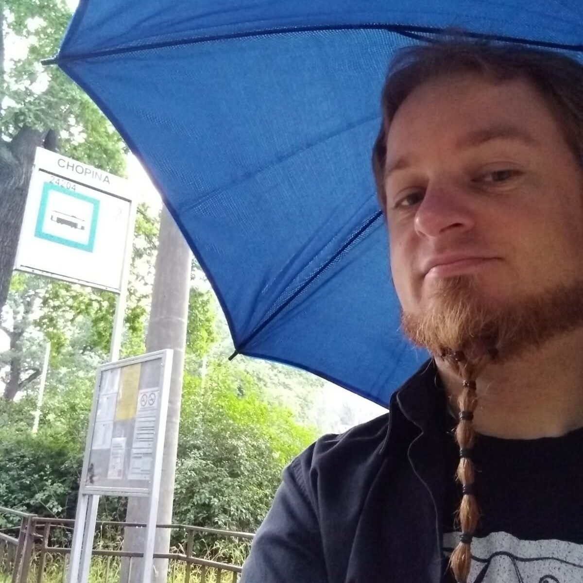 Młody mężczyzna z długą brodą robi sobie zdjęcie pod niebieskim parasolem. W tle widać przystanek autobusowy „Chopina” oraz zieleń.
